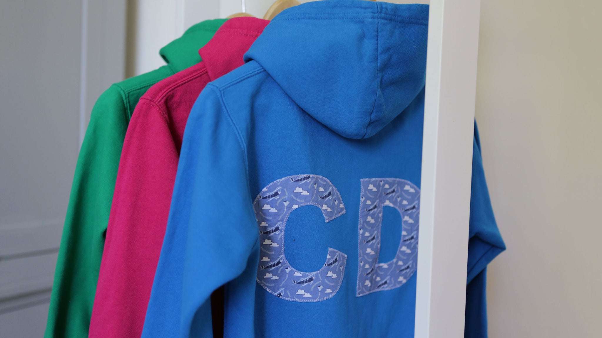 A green, pink and blue hoodie hanging on a rail. Just in view, on the back of the blue hoodie, are the letters CD appliqued on in a blue fabric with planes and clouds printed on.