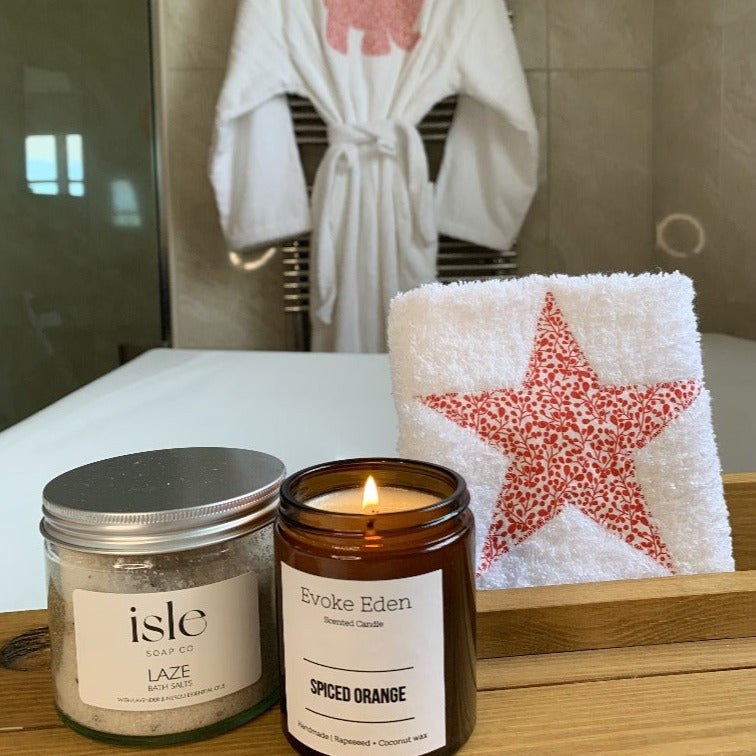 Pure white robe with an Elephant appliqued onto the back in a red berry Liberty Print. In the foreground is a face cloth with a star appliqued in the same fabric and a jar of Laze Bath Salts by Isle Soap Co and a candle in an amber glas jar in Spiced Orange from Evoke Eden